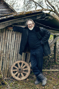 Elderly man with a beard stands in the village near a wooden shed in a sheepskin coat and glasses
