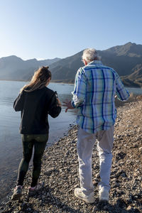 Senior man walking with his granddaughter along the lake shore with mountains.