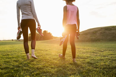 Low section of mother and daughter holding sports shoes while walking on grass at park during sunset