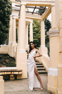A beautiful brunette lady in an elegant wedding dress poses among the columns in the old city park