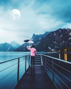 Rear view of young man with umbrella standing on pier by lake against cloudy sky