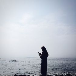Woman using mobile phone while standing on shore at beach against sky