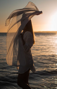 Woman holding scarf while standing at beach during sunset