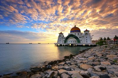 Mosque by sea against cloudy sky during sunset