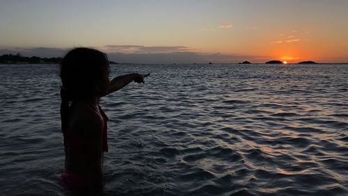 Silhouette girl pointing at sun while standing in sea during sunset