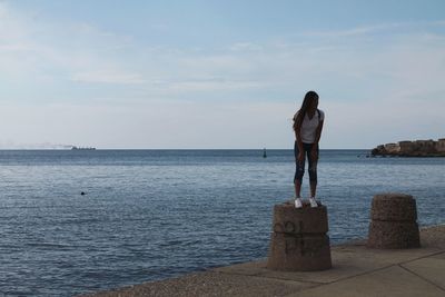 Young woman standing on bollard by sea against sky