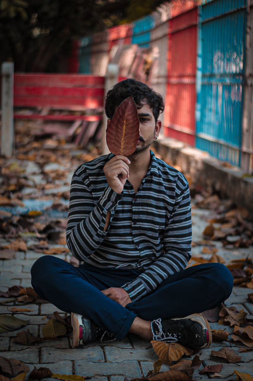 sitting, one person, adult, young adult, full length, casual clothing, lifestyles, relaxation, men, architecture, clothing, spring, person, autumn, city, leisure activity, fashion, blue, contemplation, footwear, emotion, outdoors, nature, front view, sadness, looking, photo shoot, arts culture and entertainment, street, homelessness