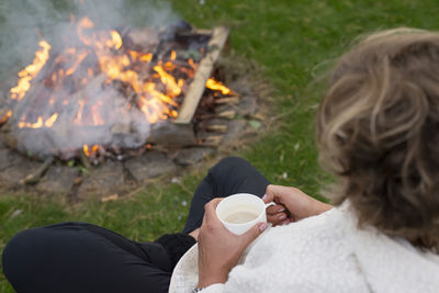 High angle view of woman holding coffee cup sitting by campfire at lawn
