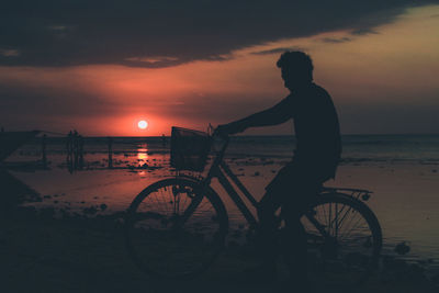 Silhouette man riding bicycle on beach against sky during sunset