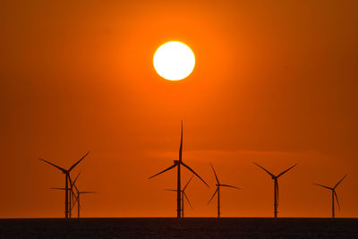 Silhouette wind turbines in sea against sky during sunset