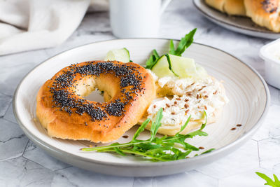 Bagel sandwich with poppy seeds with ricotta, cucumber and arugula on a plate on the table. 