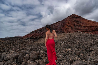 Rear view of woman wearing red dress standing on rocks against cloudy sky