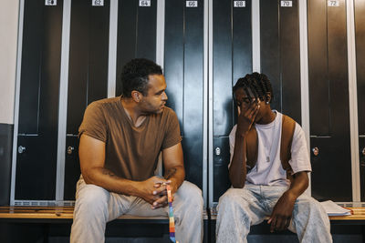 Male sports coach consoling sad teenage boy sitting on bench in locker room at junior high school