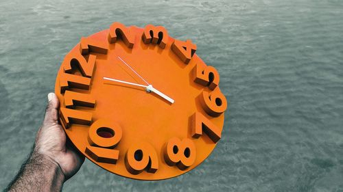 Cropped hand holding orange clock by sea