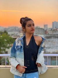 Young woman looking away while standing by railing against sunset sky