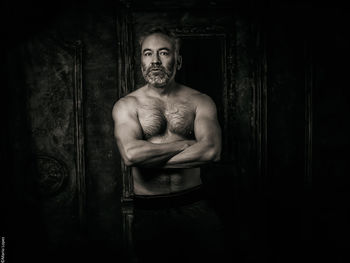 Shirtless man standing against wall arms crossed 