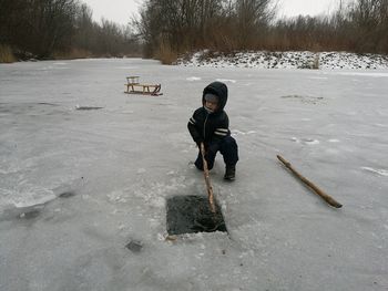 Boy holding stick in water while kneeling on ice field during winter