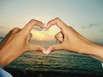 Cropped hands making heart shape at beach against sky during sunset