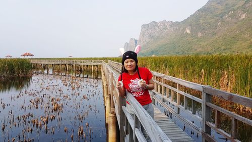 Portrait of smiling mature woman standing on footbridge over lake against clear sky