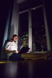 Freelance man in glasses working, sits at the window on the floor in front of a skyscraper at night