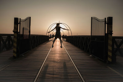 Full length of silhouette teenage girl jumping on footbridge against sky during sunset
