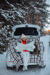 A young blonde girl sits in the trunk of a white car in winter with a paper map in her hands