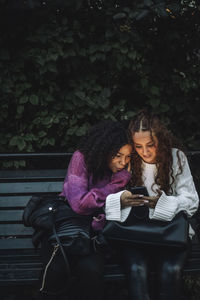 Curious female friends sharing smart phone while sitting on bench near plants