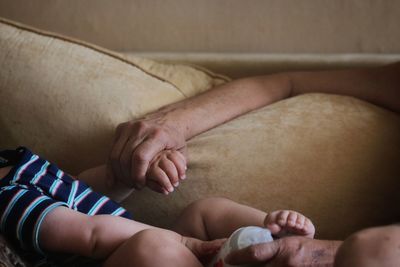 Cropped image of father holding hands with baby on sofa at home