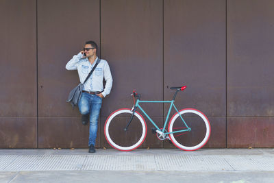 Man talking on phone while standing with bicycle against wall
