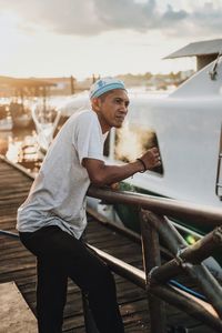 Side view of young man standing on railing against sky