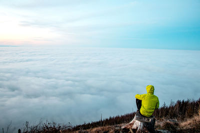Young hiker in green jacket sits on a tree stump and enjoys the view of the gray-blue sea of clouds 