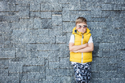 Boy wearing sunglasses with arms crossed standing against stone wall