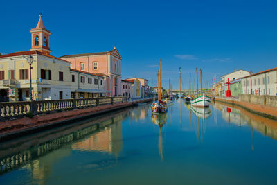 Boats moored in canal by buildings against blue sky