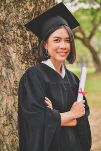 Portrait of young woman in graduation gown holding certificate while standing by tree at park