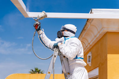 Anonymous employee in protective uniform and mask standing on ladder and using air brush to paint frame structure against cloudy sky on sunny day