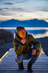 Full length of young man crouching on boardwalk against sky during sunset