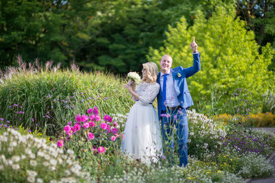 Groom gesturing while bride holding bouquet while standing amidst plants at park