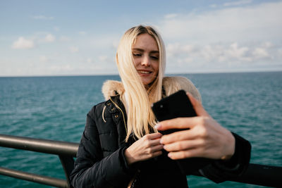 A beautiful smiling blonde girl makes a selfie on the phone against the background of the sea. 