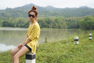 Portrait of woman wearing sunglasses while sitting on bollard at lakeshore
