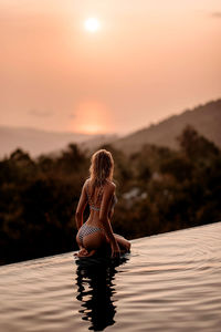 Side view of woman sitting on the edge of the poolagainst sky during sunset