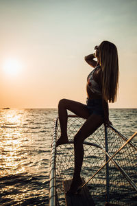 Woman sitting by sea against sky during sunset