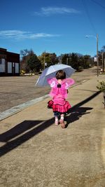 Rear view of girl in angel costume holding umbrella while walking on sidewalk