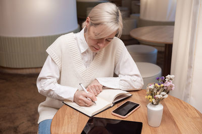 A cute girl is sitting in a cafe and making notes in a diary, there is a phone and a tablet nearby