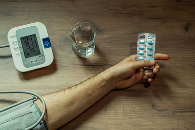 A man's hand with a pill in his hand, a blood pressure monitor and a glass of water on a wooden