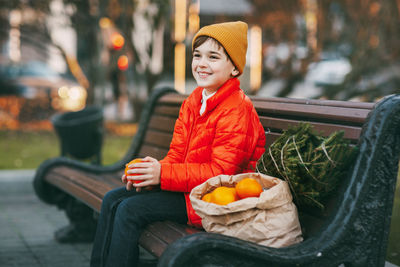 A boy in a bright orange jacket and a fashionable yellow hat sits on a bench, laughs, throws oranges 