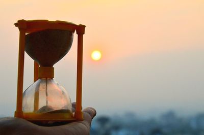 Close-up of hand holding hourglass against sky during sunset