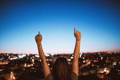 Rear view of woman pointing with arms raised against clear sky at dusk