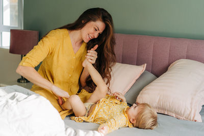 Young red-haired mother plays and tickles her little cheerful daughter sitting on the bed.