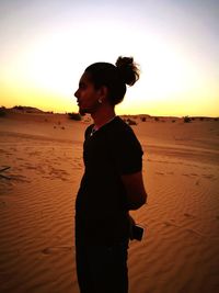 Side view of woman standing at beach against sky during sunset