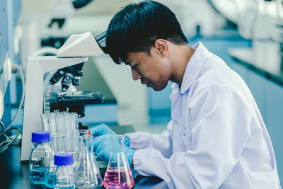 Asian man lab technician in protective glasses and gloves sits next to a microscope in laboratory.